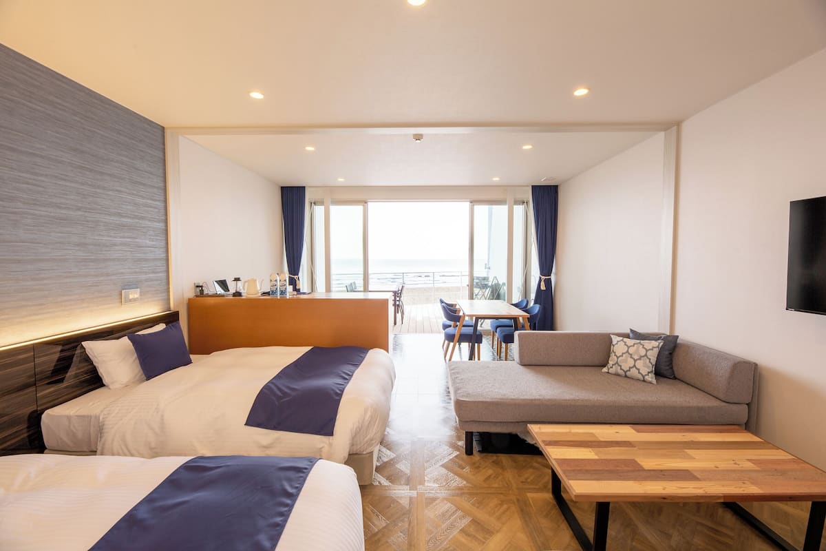 A hotel room with two beds, a sofa, a wooden table, and a view of the sea.