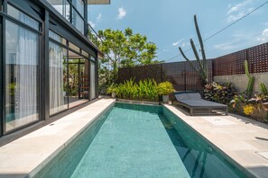 A modern outdoor pool area with a clear blue pool, a wooden deck, and a cactus plant. - Villa Greta (Canggu)