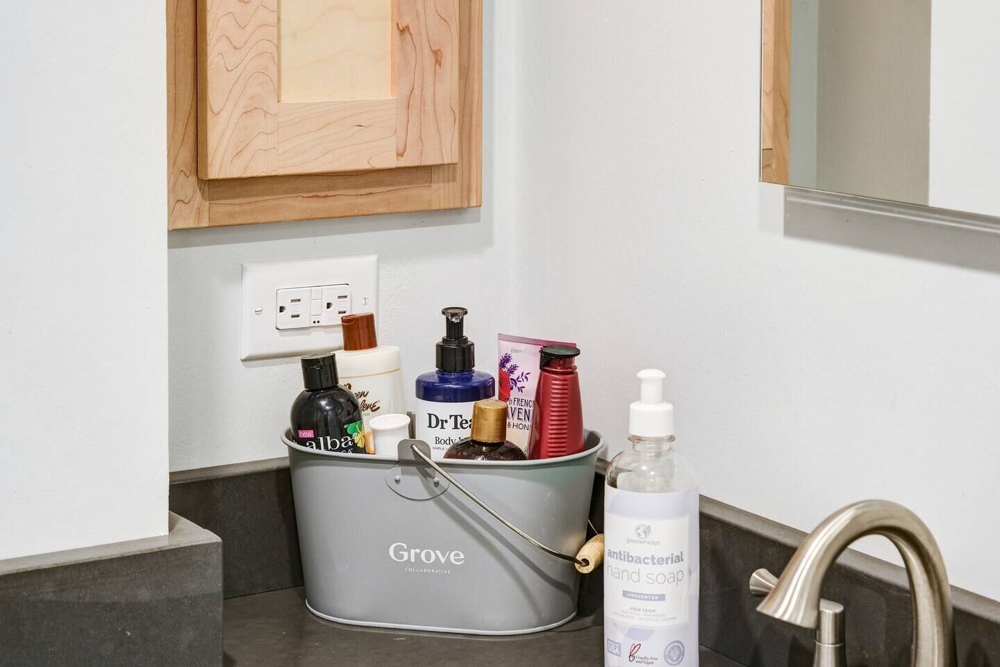 A bathroom with a grey basin containing toiletries, a bottle of antibacterial hand soap, and a mirror above the sink.