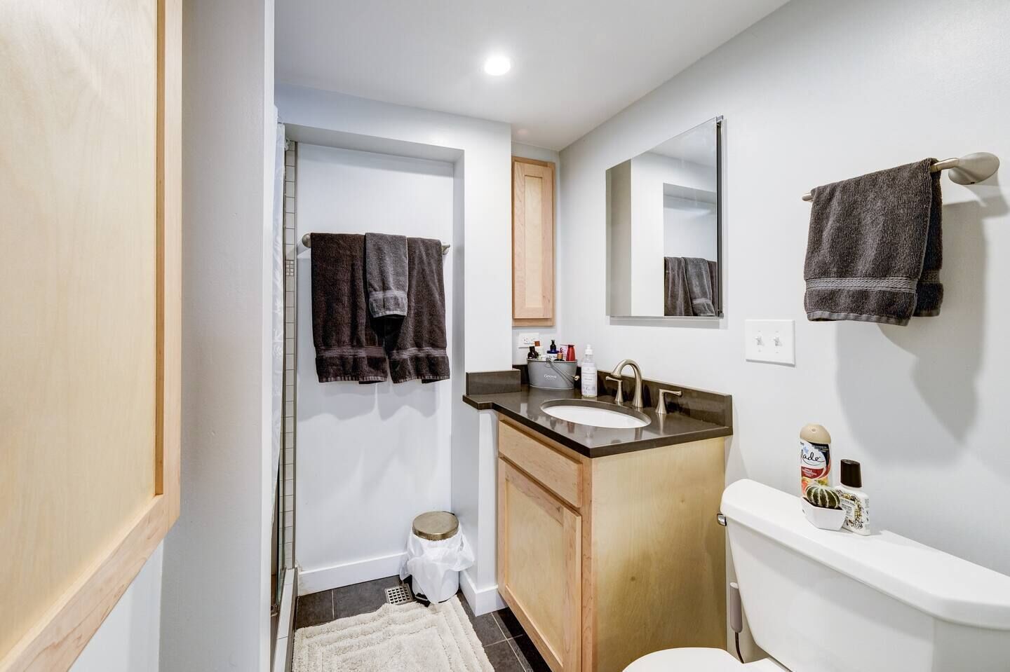 A compact bathroom with a wooden vanity, a white sink, and a toilet. There are two towels hanging on the wall, and a small step stool is visible.