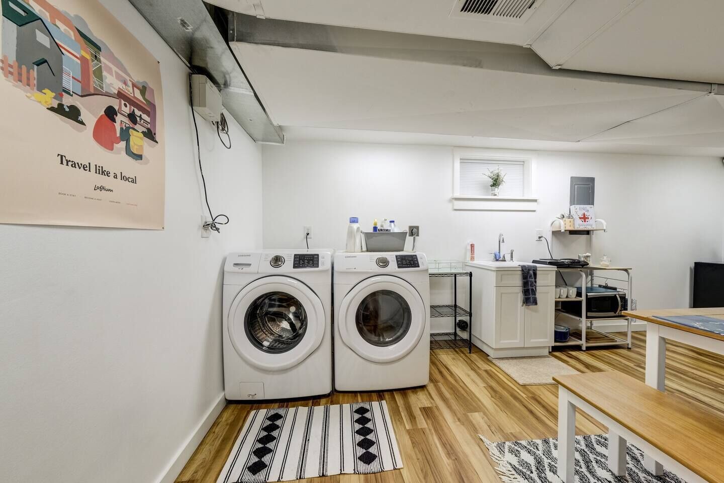 A laundry room with two front-loading washing machines, a sink, a small table, and a bench.