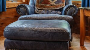 A corner room with a leather sofa and ottoman, a wooden chair, a hat on the wall, and a view of a forest through large windows. - Heartland Lodge: Multi-fam cabin in Hocking Hills (Logan)