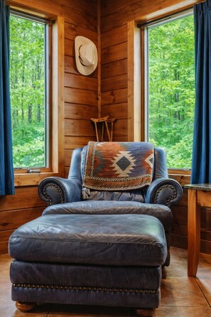 Ein Eckzimmer mit einer Ledersofa und Hocker, einem Holzstuhl, einem Hut an der Wand und Blick auf einen Wald durch große Fenster.
