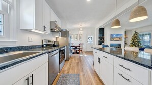 Cocina moderna con armarios blancos, encimeras de granito y electrodomésticos de acero inoxidable. Hay un comedor con mesa y sillas, y un árbol de Navidad al fondo.