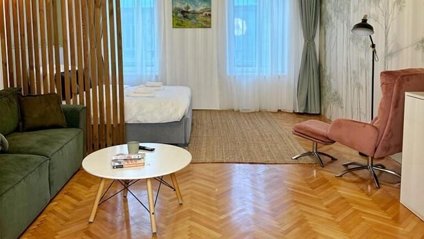 Habitació moderna amb sòl de fusta, un sofà verd, un llit amb roba de llit blanca, una taula de cafè rodona i una cadira rosa.
