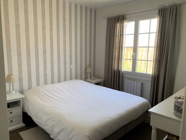 Una cama bien hecha en una habitación con papel tapiz a rayas, una mesita de noche con lámpara y una ventana con cortinas.