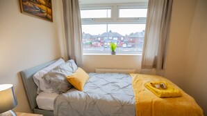 A bedroom with a bed, a bedside table with a lamp, a window with a view of buildings, and a potted plant on the windowsill. - 3bed House in Stockton-on-tees- Free Wifi, Parking (Stockton-on-Tees)