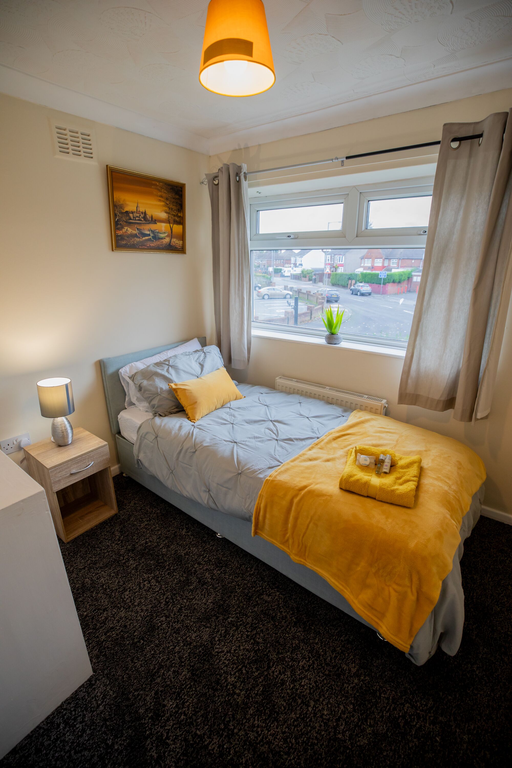 A single bed with a headboard, a bedside table with a lamp, a window with curtains, and a painting on the wall.