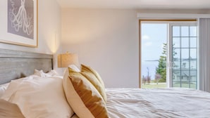 A hotel room with a bed, a lamp, a framed picture, and a view of the sea through a large window.