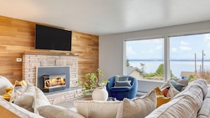 A living room with a fireplace, a television, and a large window with a view of the sea.