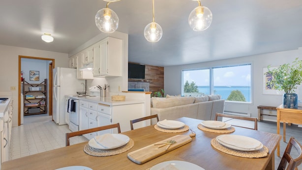A modern kitchen with white cabinets, a dining area with a wooden table set for four, and a large window offering a view of the sea.