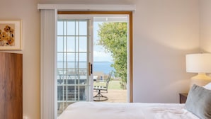 A bedroom with a bed, a nightstand, a lamp, a sliding glass door leading to an outdoor patio with a table and chairs, and a view of the sea.