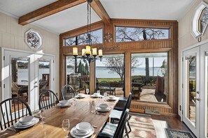 A dining area with a wooden table set for six, large windows offering a view of the outdoors, and a chandelier above. - Bear Creek Lodge by Avantstay Lakefront + Dock (Mooresville)