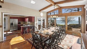 A spacious dining area with a wooden table set for six, surrounded by chairs. A kitchen with wooden cabinets and a refrigerator is visible in the background. - Bear Creek Lodge by Avantstay Lakefront + Dock (Mooresville)