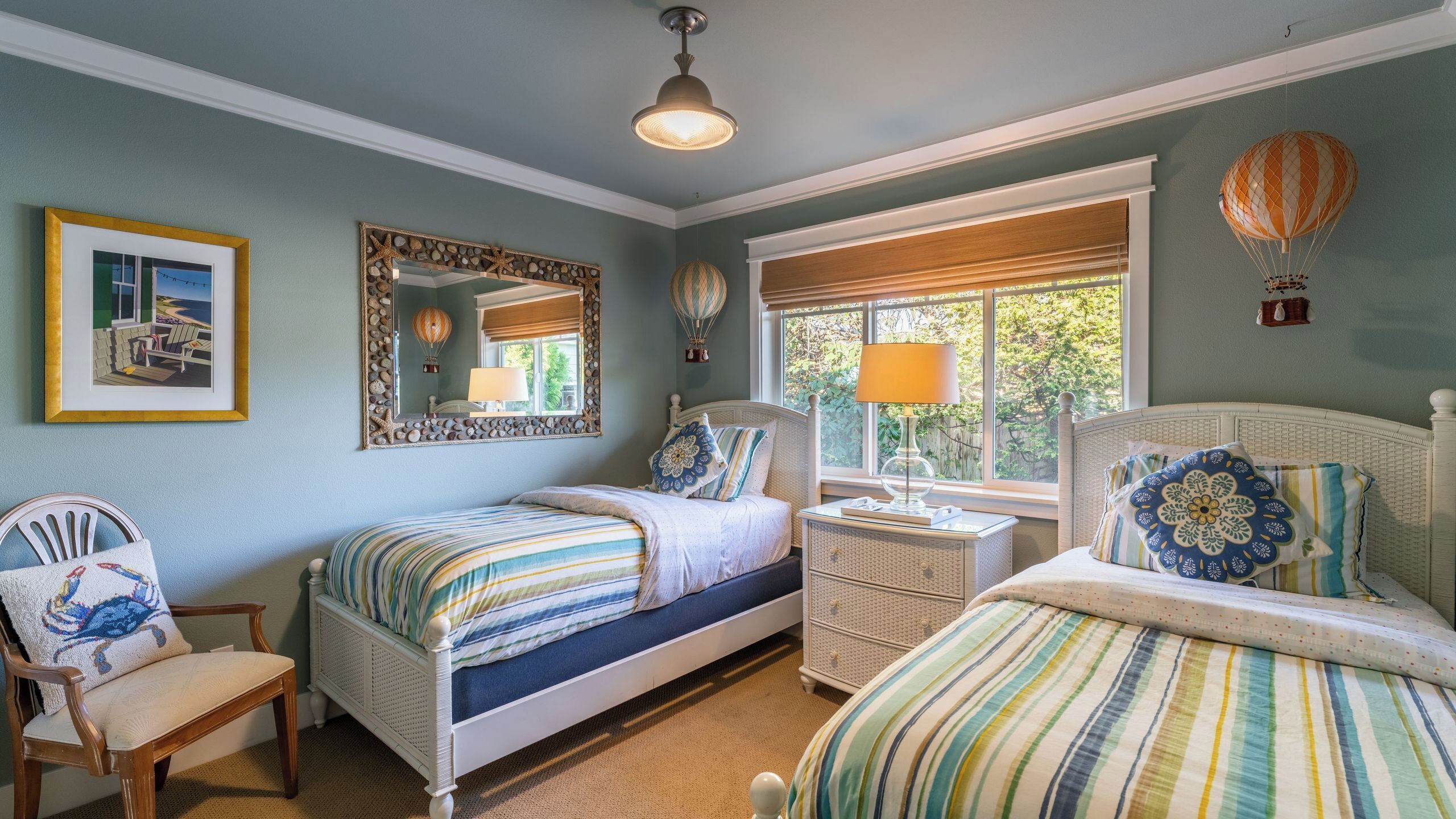 A bedroom with two beds, a window with a view, a mirror with a decorative frame, and a ceiling fan.