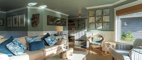 A cozy living room with a sofa, armchairs, and a coffee table. There are framed pictures on the walls and a window with a view.