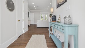 A hallway with a white door, a white sofa, a light blue console table with decorative items, and a white rug on a wooden floor. - Yeah, Bay-by! Modern Farmhouse in Old Town Bay Saint Louis (Bay St. Louis)