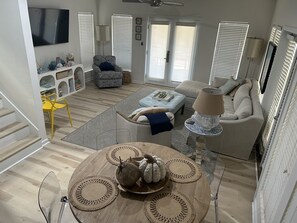 Een woonkamer met een bank, fauteuils, een salontafel en een televisie. Er zijn ramen met zonwering, een lamp en een trap.