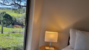 A bedroom with a bed, a bedside table with a lamp, and a view of a green landscape through a window.
