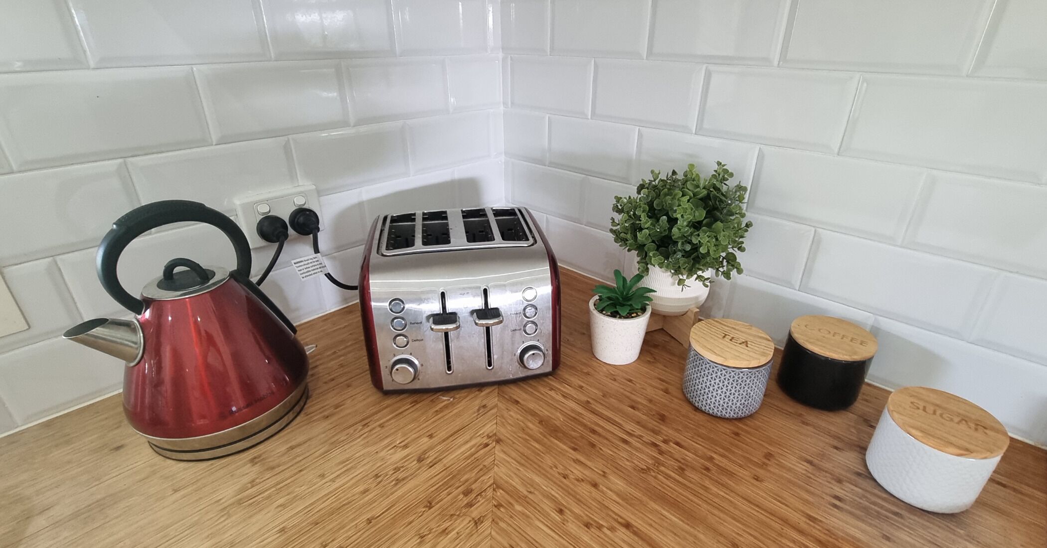 A kitchen counter with a red kettle, a silver toaster, a small potted plant, and two containers labeled "TEA" and "COFFEE".