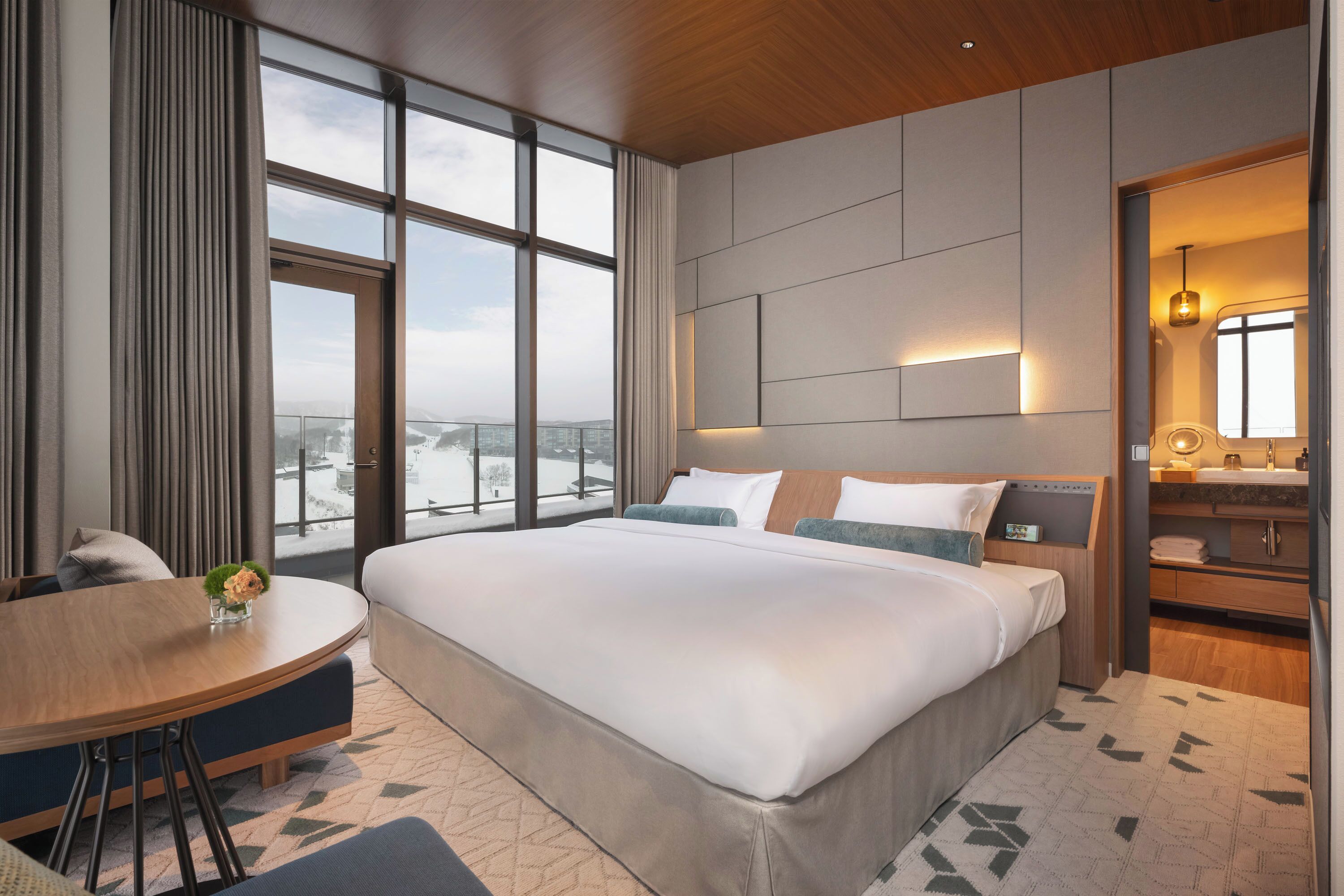 Habitación de hotel moderna con una cama grande, una mesa de comedor redonda, un baño con tocador y vistas de la ciudad a través de amplias ventanas.