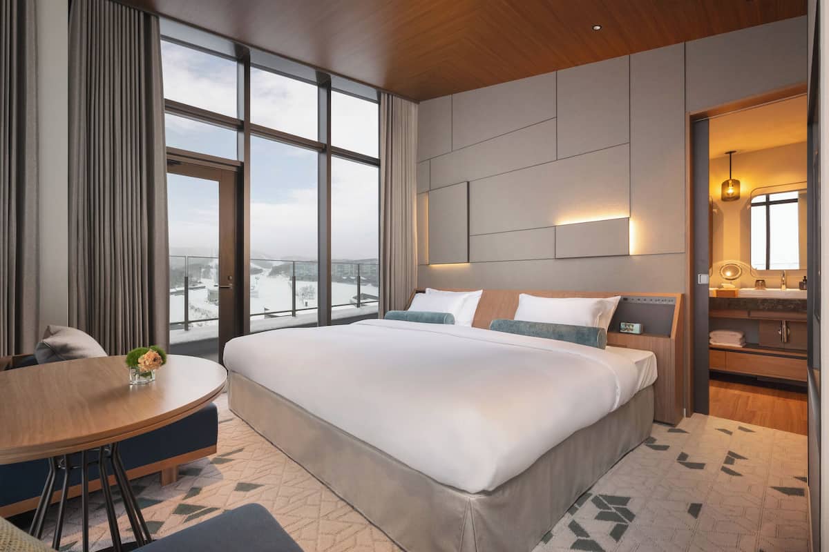 Ein modernes Hotelzimmer mit einem großen Bett, einem runden Esstisch, einem Badezimmer mit Waschtisch und Blick auf die Stadt durch große Fenster.