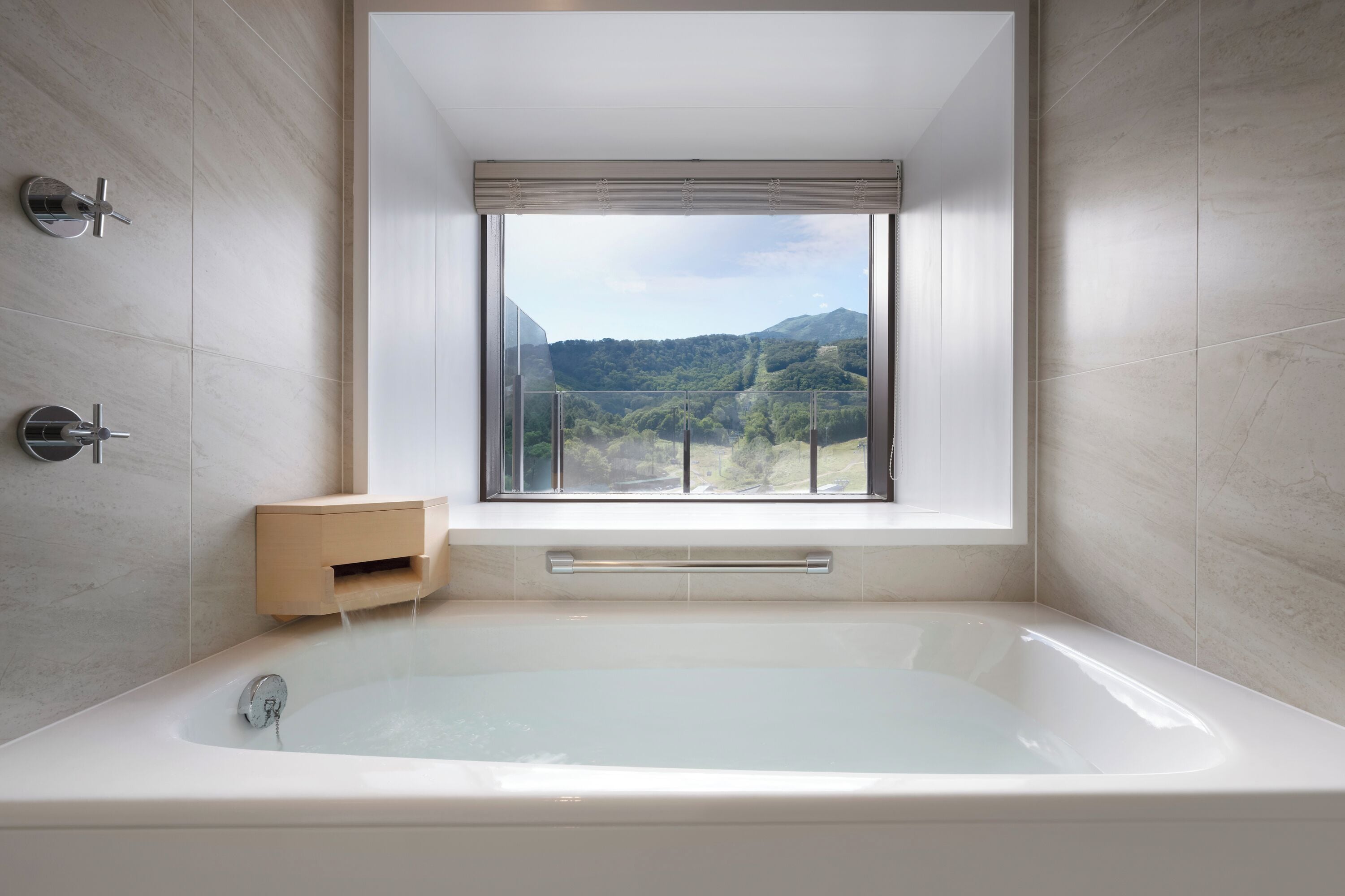 A modern bathroom with a large bathtub, a window with a view of green hills, and a small wooden side table.