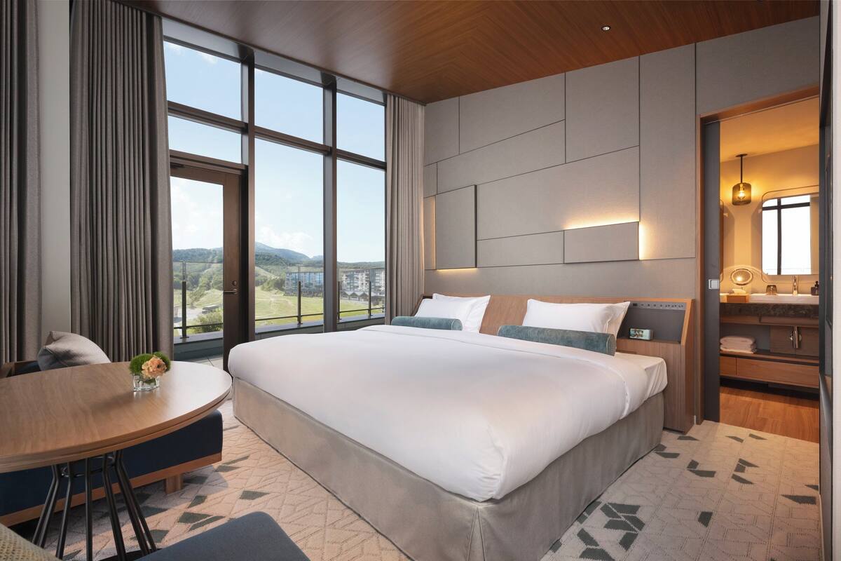 Ein modernes Hotelzimmer mit einem großen Bett, einem runden Esstisch und Blick ins Freie.