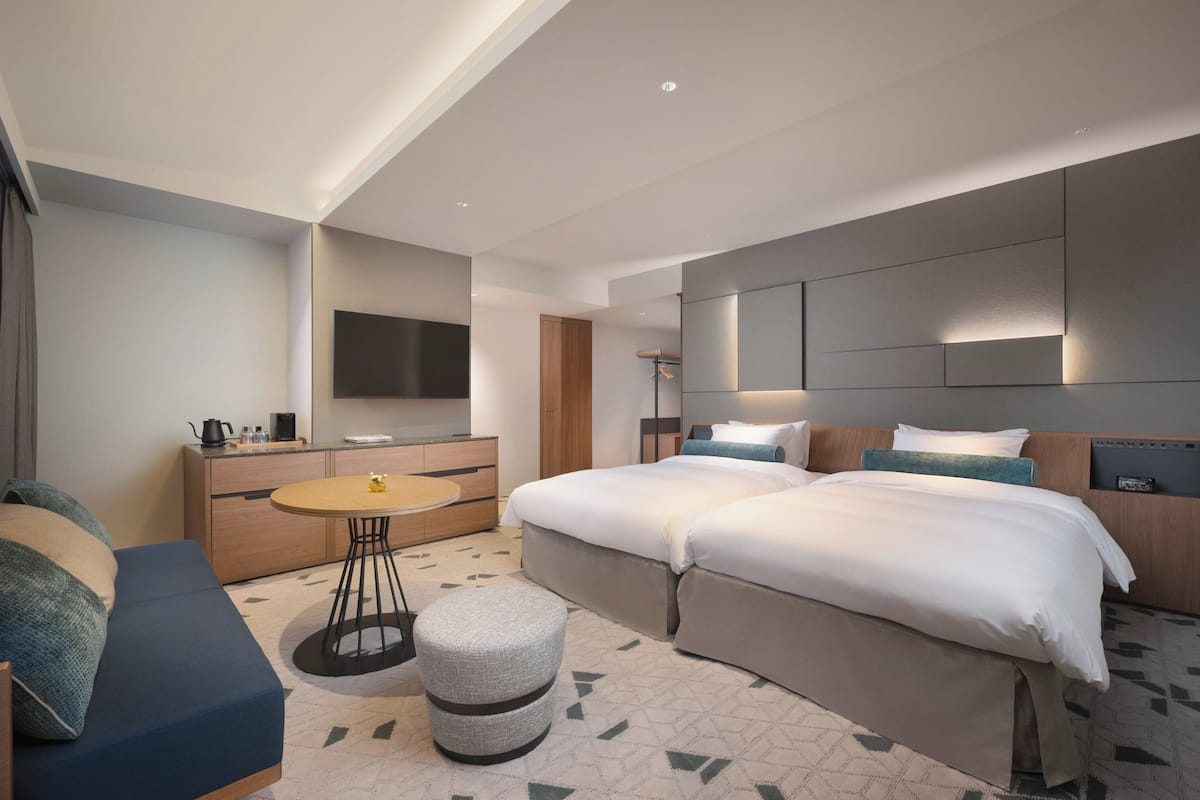 Ein modernes Hotelzimmer mit einer Couch, einem runden Esstisch, einem kleinen Hocker, einem Doppelbett, einem Fernseher und einem integrierten Bettkopfteil mit Beleuchtung.