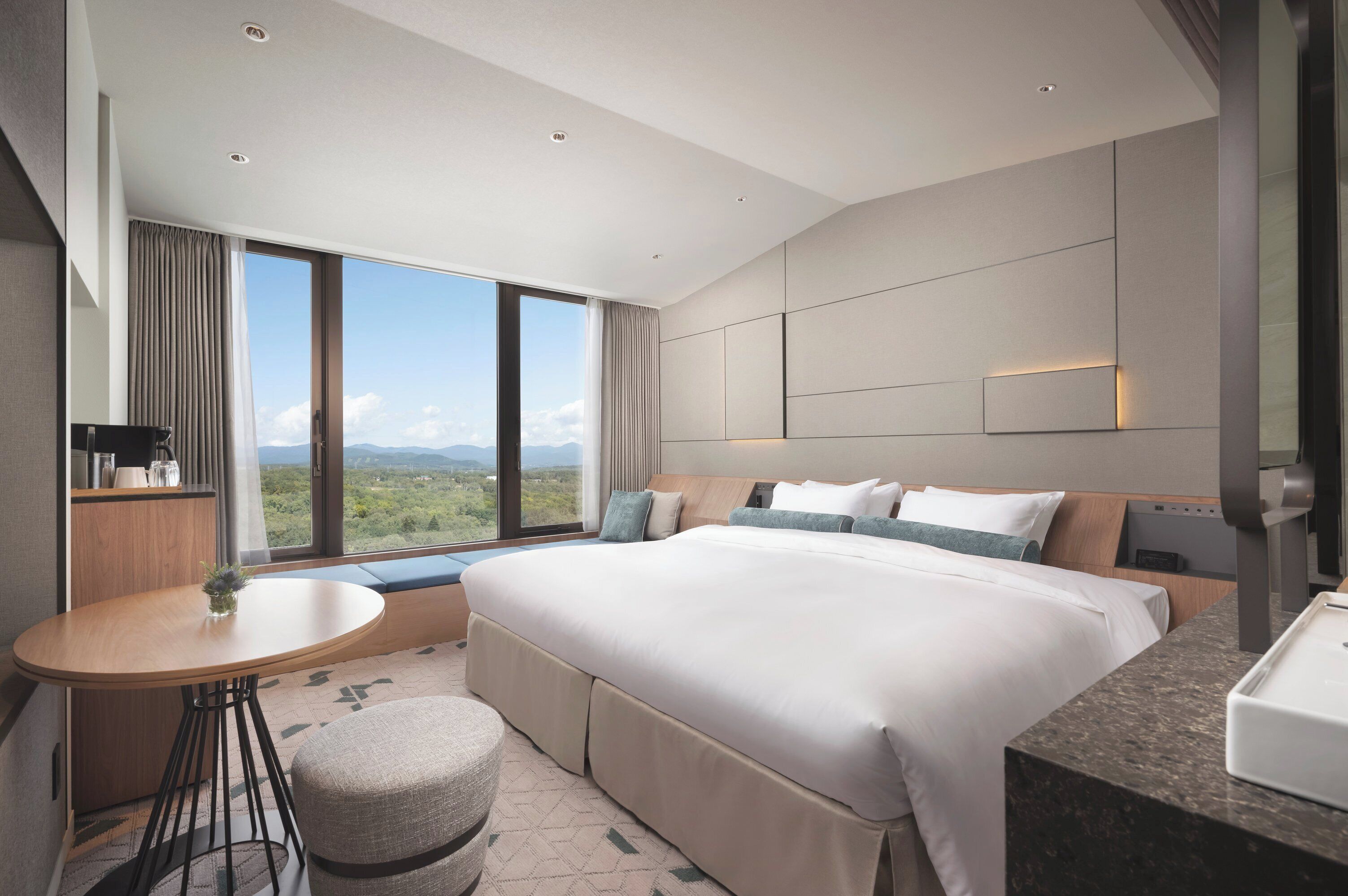 A modern hotel room with a large bed, a small round table, a seating area, and a view of the outdoors.