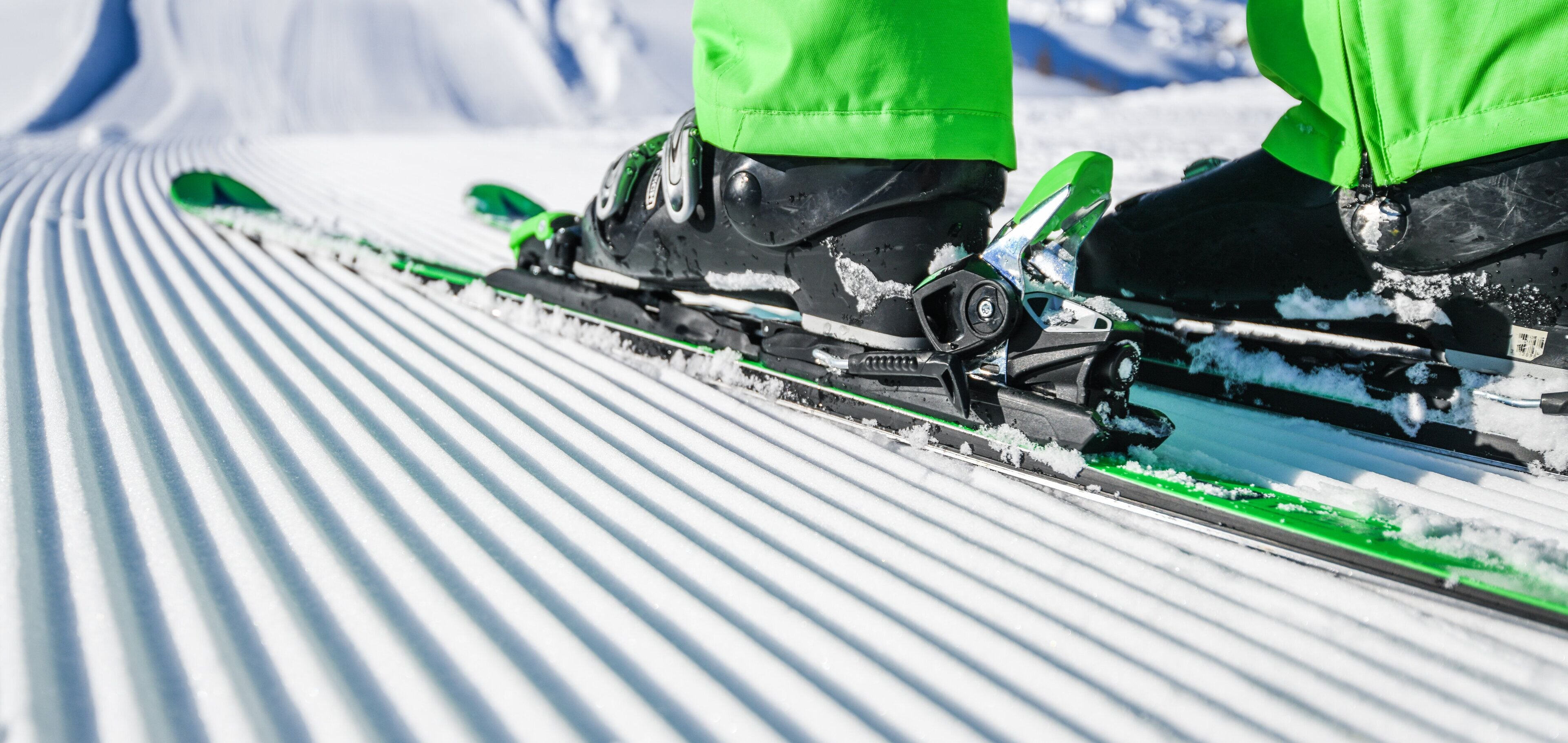 A close-up of a ski track with green and black ski boots and bindings.