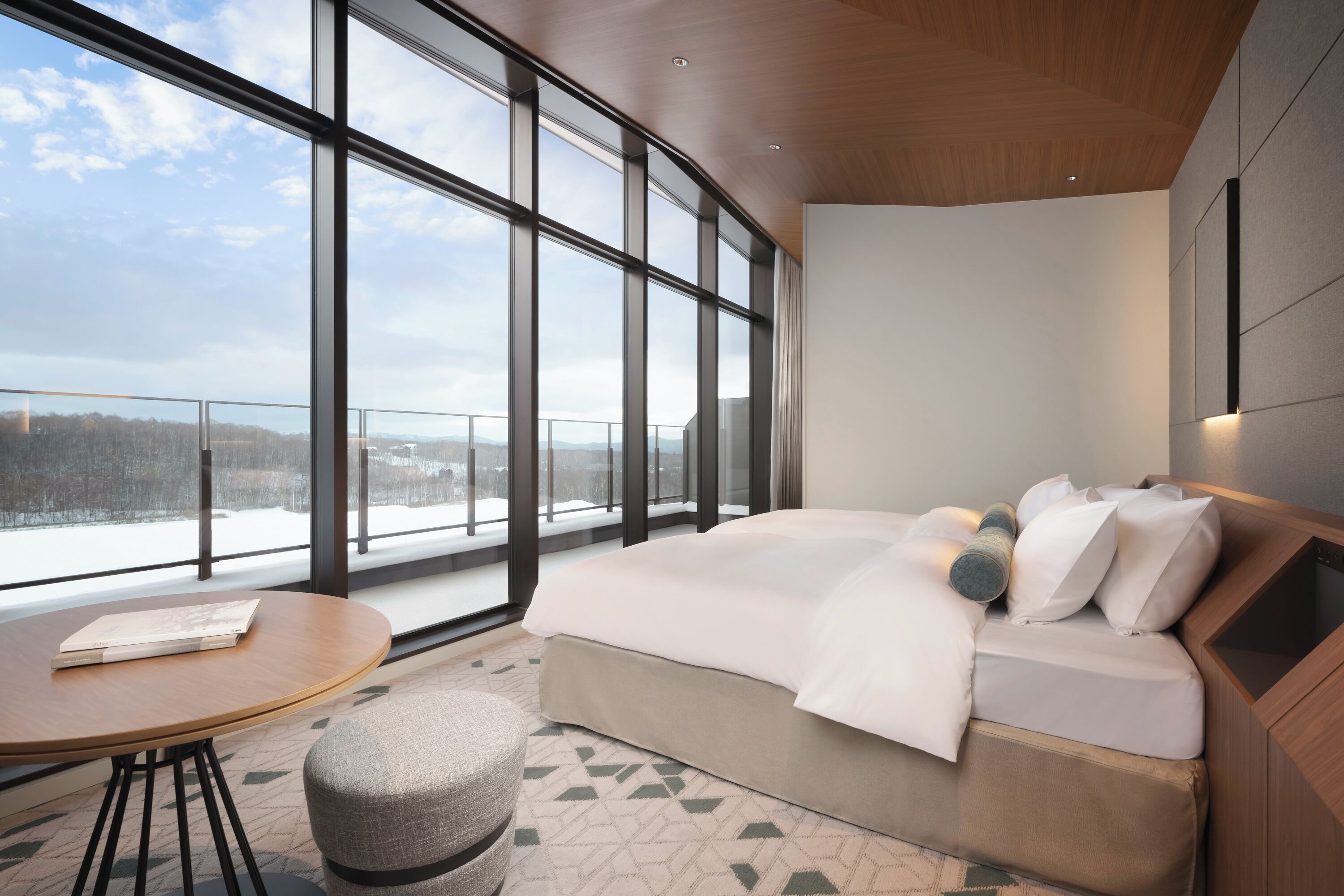 Una habitación de hotel moderna con un ventanal grande, una cama, una mesa redonda y un taburete pequeño.