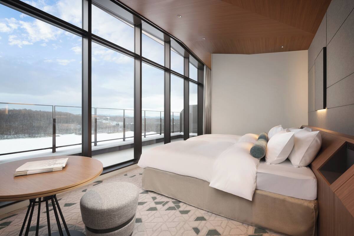 Ein modernes Hotelzimmer mit großem Fenster, einem Bett, einem runden Tisch und einem kleinen Hocker.