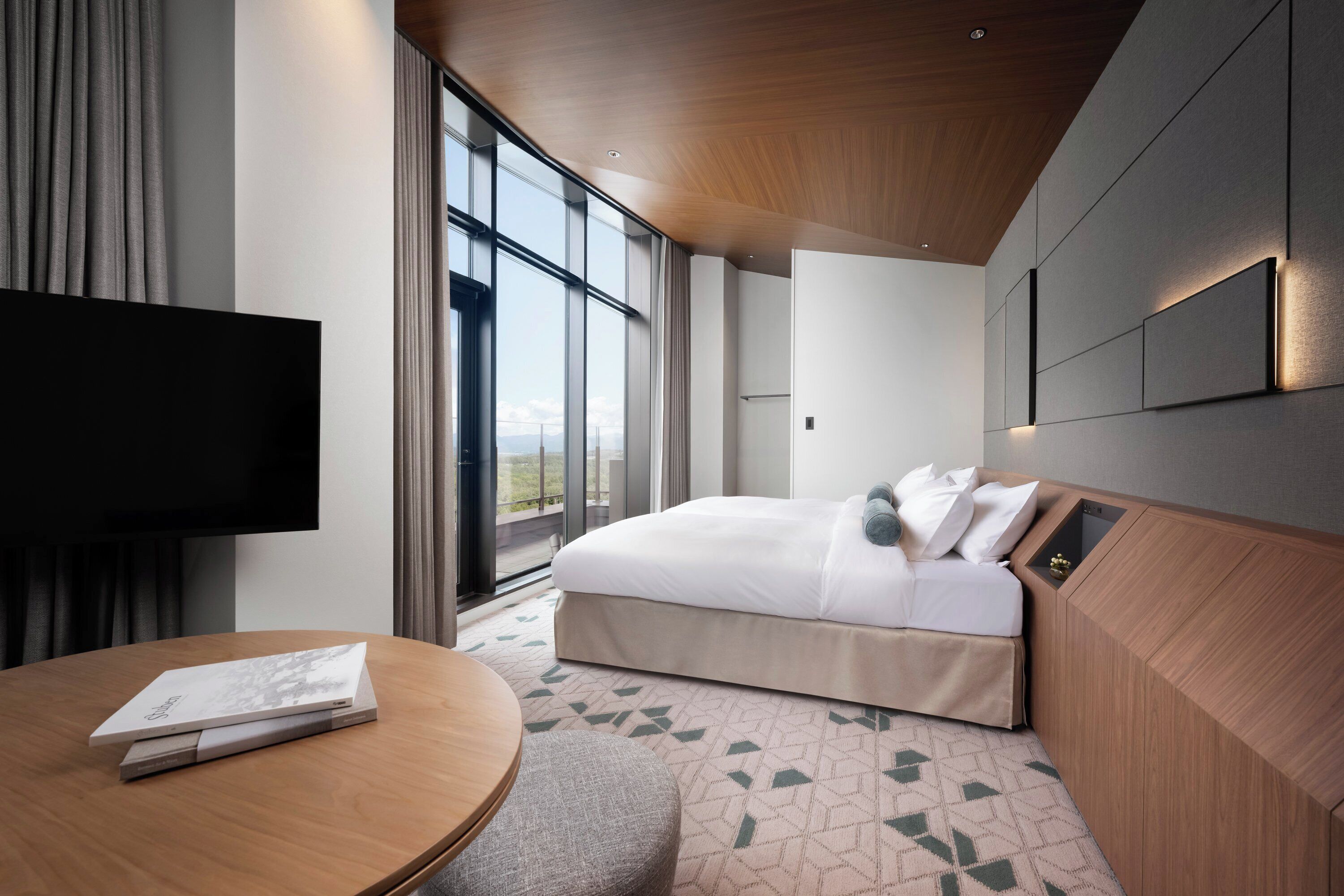 Una habitación de hotel moderna con una cama grande, un televisor de pantalla plana, una mesa redonda con un libro y un ventanal con vistas a la ciudad.