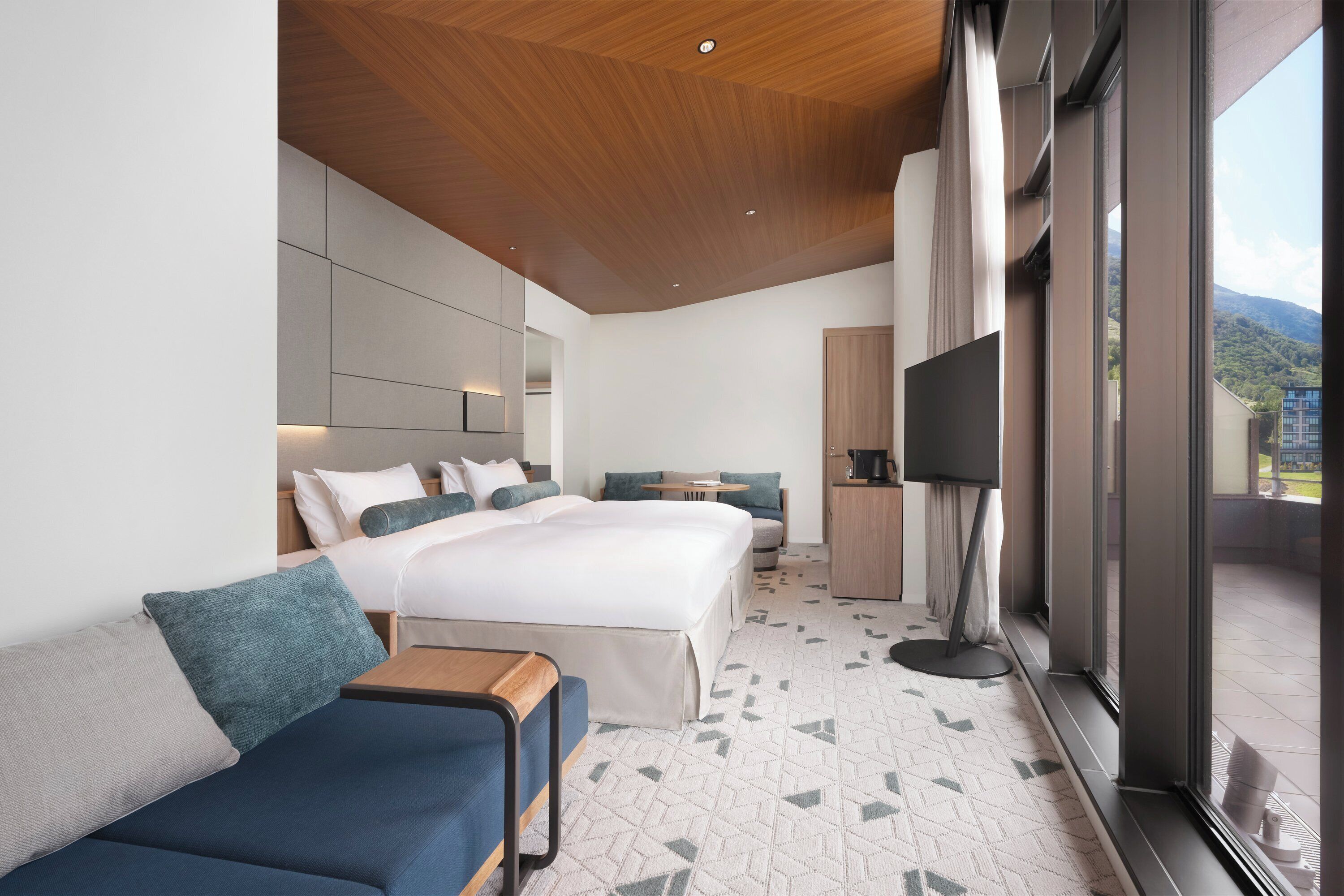 A modern hotel room with a large bed, a sofa, a TV, and a balcony with a mountain view.