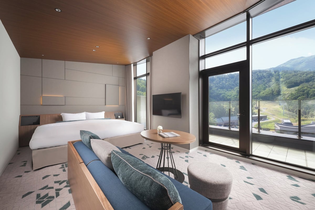Ein modernes Hotelzimmer mit einem großen Bett, einer Couch, einem kleinen Tisch und Blick auf eine gebirgige Landschaft durch große Fenster.