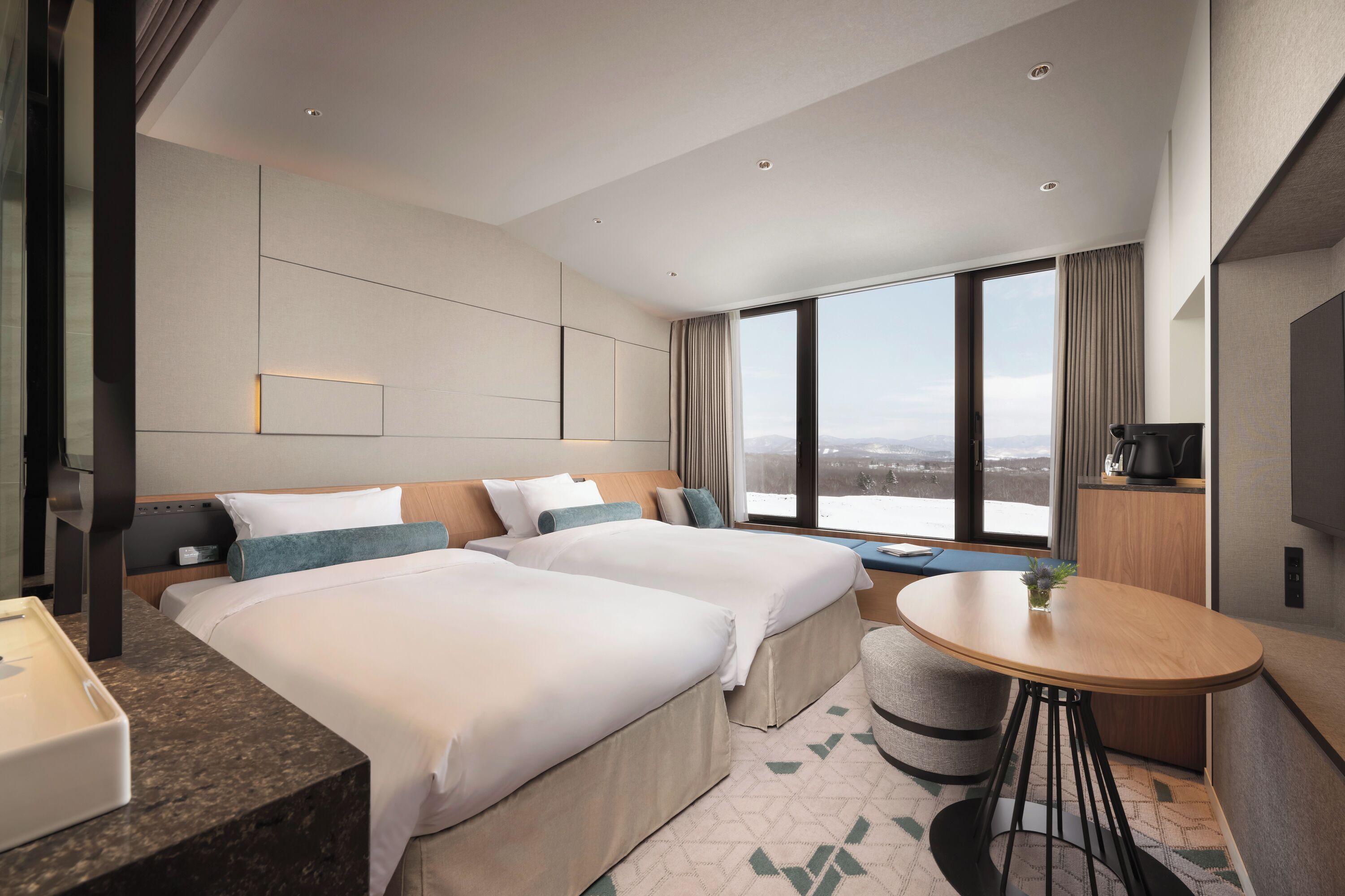 Habitación de hotel con dos camas, una mesita, televisión y un ventanal grande con vistas a la ciudad.
