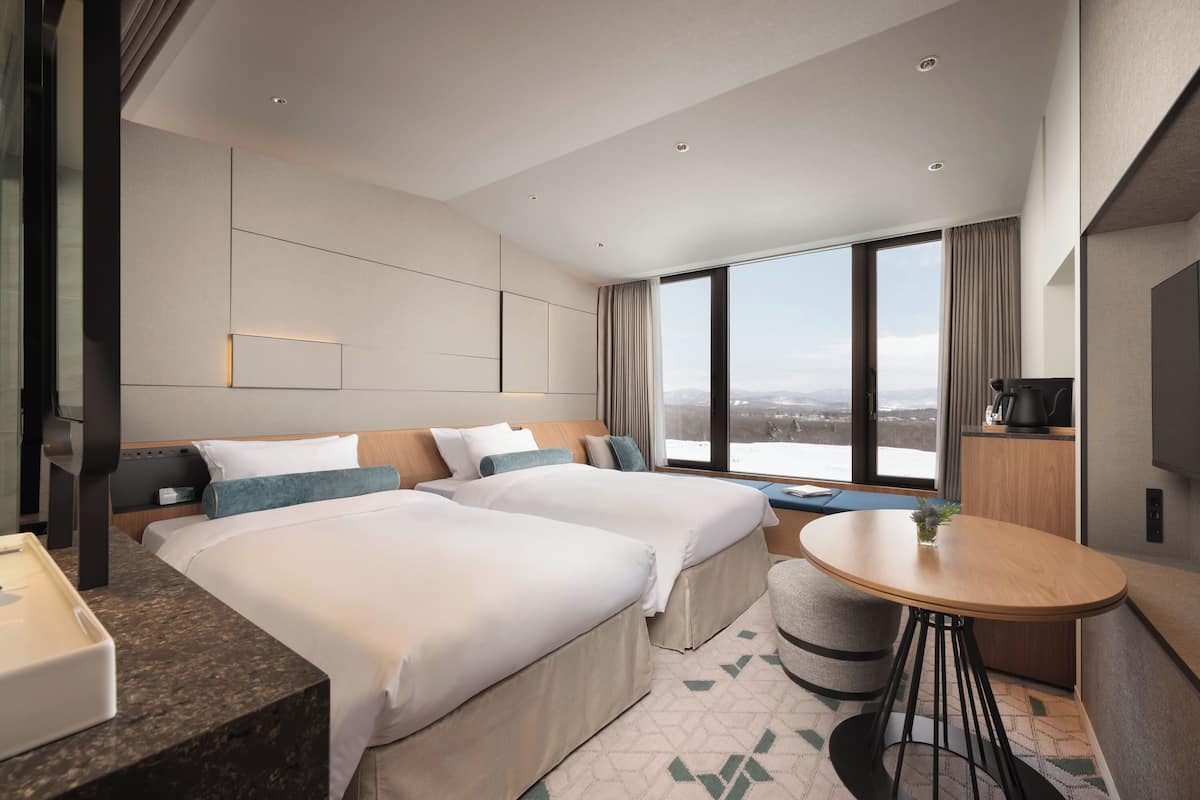 Habitación de hotel con dos camas, una mesita, televisión y un ventanal grande con vistas a la ciudad.