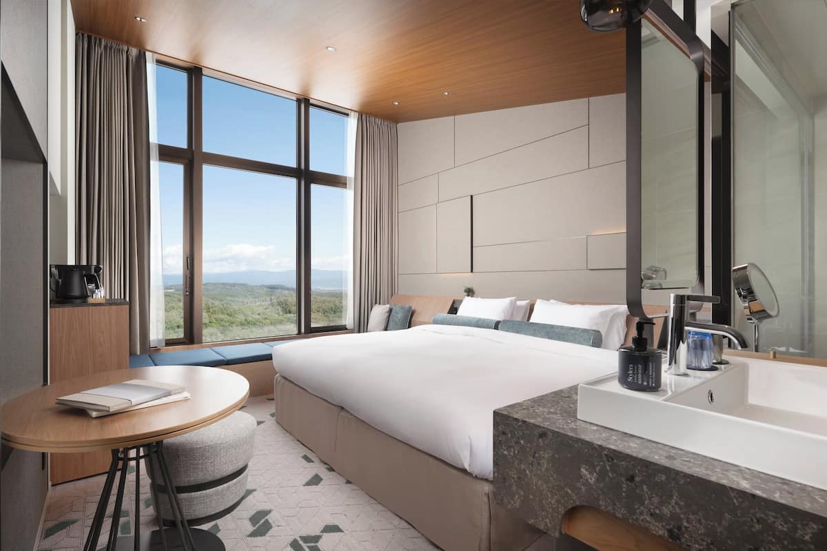 Ein modernes Hotelzimmer mit einem großen Bett, einem runden Tisch, einem Badezimmer mit einem großen Spiegel und Blick auf die Landschaft durch große Fenster.