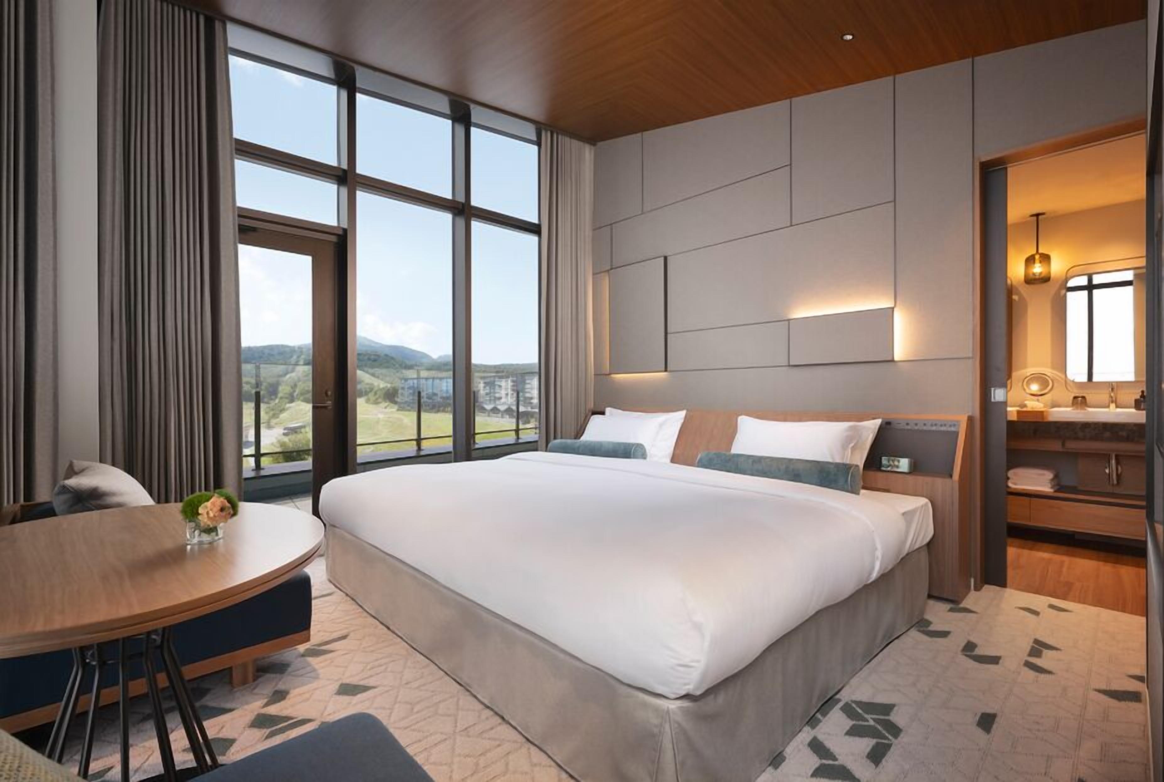 A modern hotel room with a large bed, a round dining table, and a bathroom with a mirror and sink.