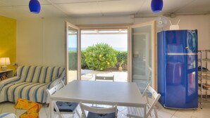 Habitació amb taula de dinar i cadires, un sofà, nevera blava, i vista a la vegetació i al mar per una porta oberta.