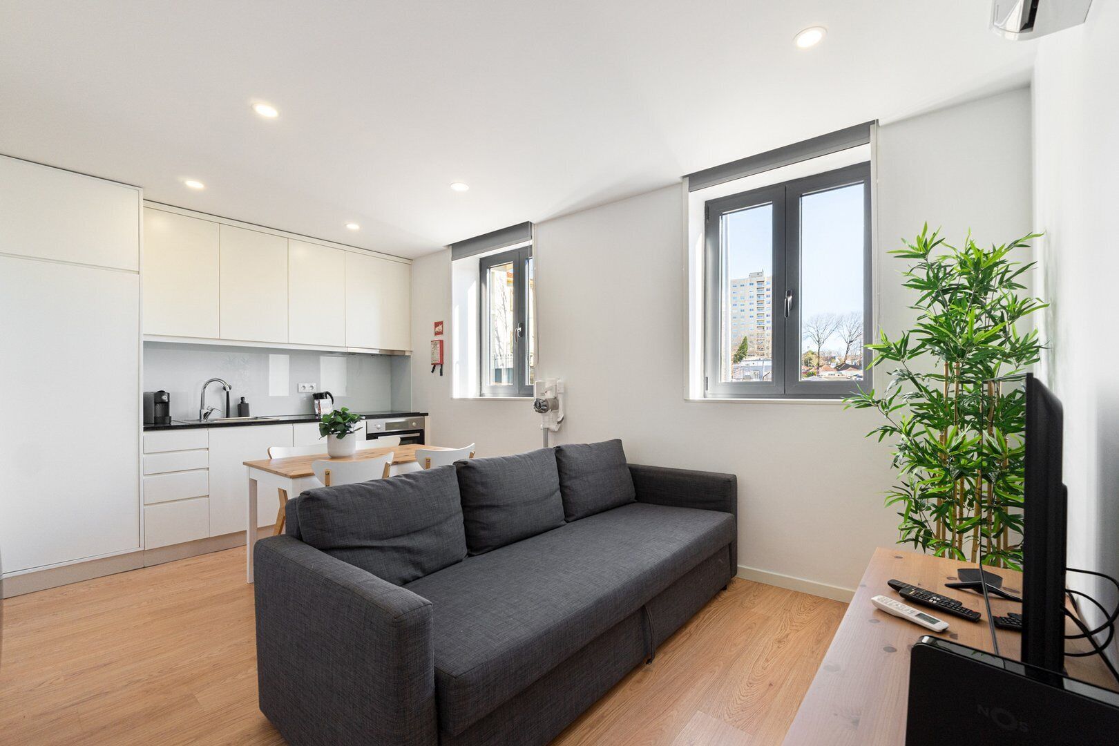 A modern apartment with a grey sofa, a dining table, and a kitchen in the background.
