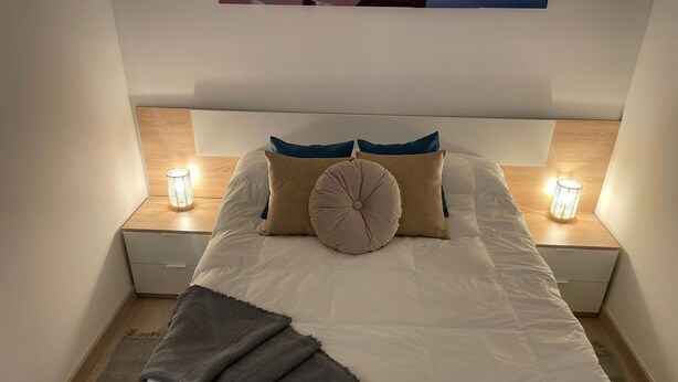 Un lit bien fait, recouvert d’une couette blanche, avec deux oreillers et un plaid gris. De chaque côté du lit, on trouve une table de chevet sur laquelle est posée une lampe.