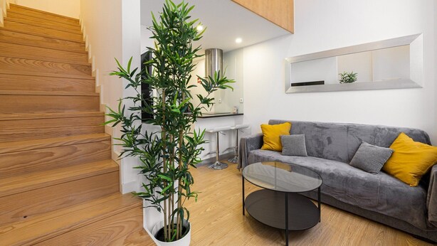 A modern living room with a staircase, a grey sofa with yellow cushions, a round glass coffee table, and a tall bamboo plant in a white planter. - GuestReady - King's Retreat (Porto)