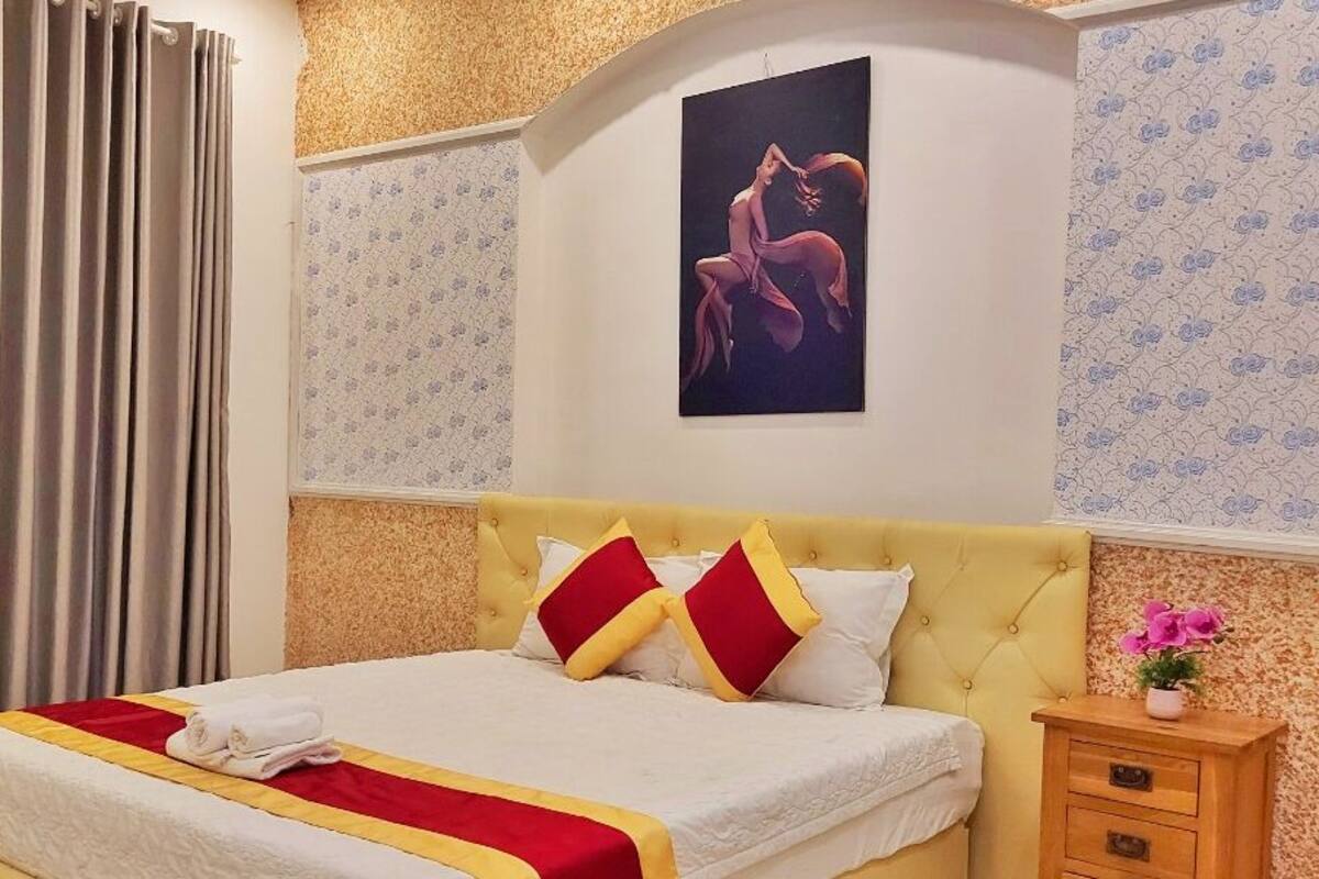 Ein ordentlich bezogenes Bett mit einer Decke in Rot und Gelb, einem gelben Kopfteil und einem einzelnen Kissen in Rot und Gelb. Dazu ein Nachttisch aus Holz mit einer Blumenvase rechts neben dem Bett.