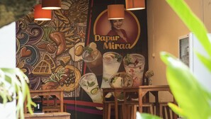 A restaurant with a mural, wooden furniture, and hanging pendant lights. - Miraa Guest House & Resto (Denpasar)