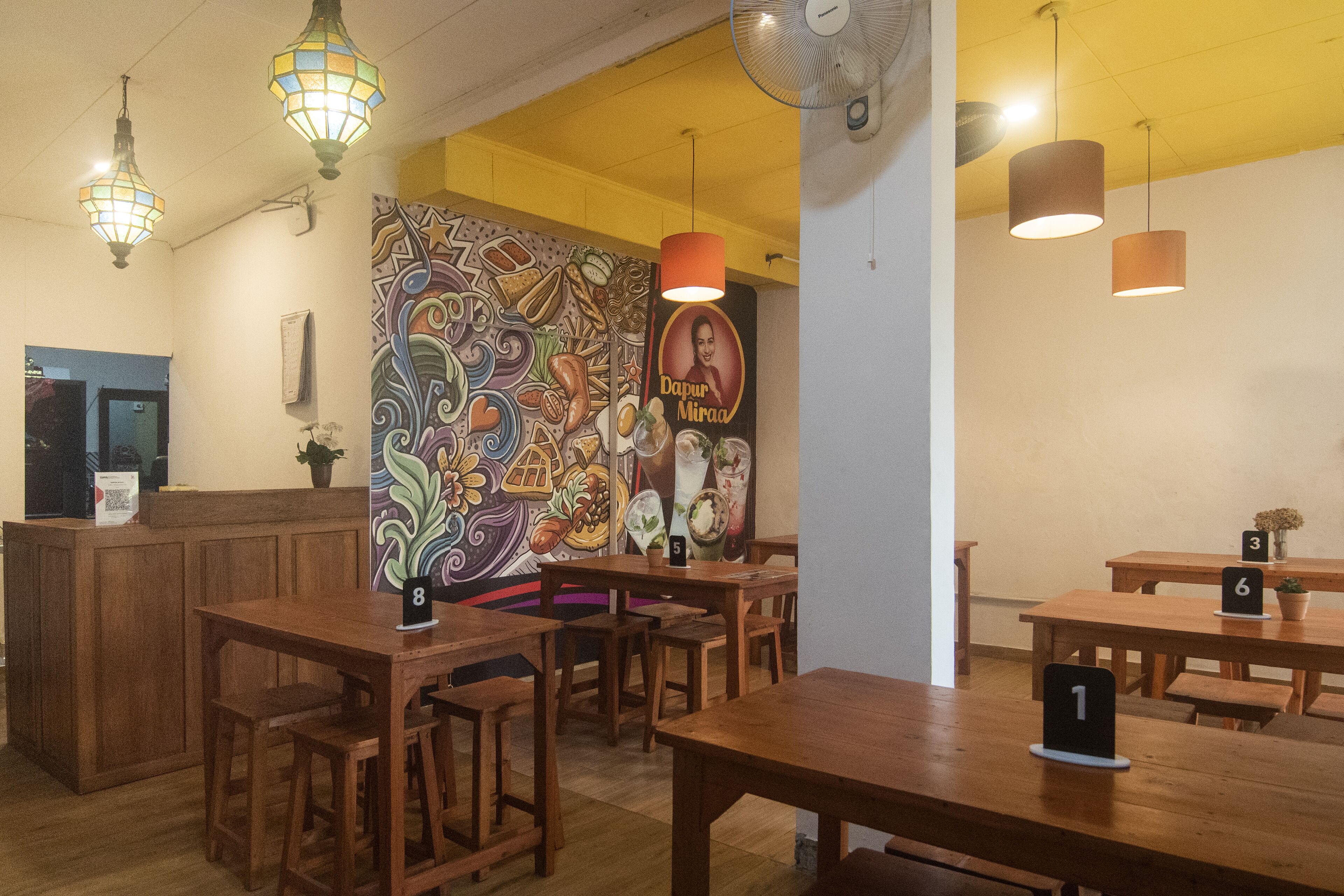 A restaurant interior with wooden tables and chairs, numbered seating, a mural on the wall, and a ceiling fan.