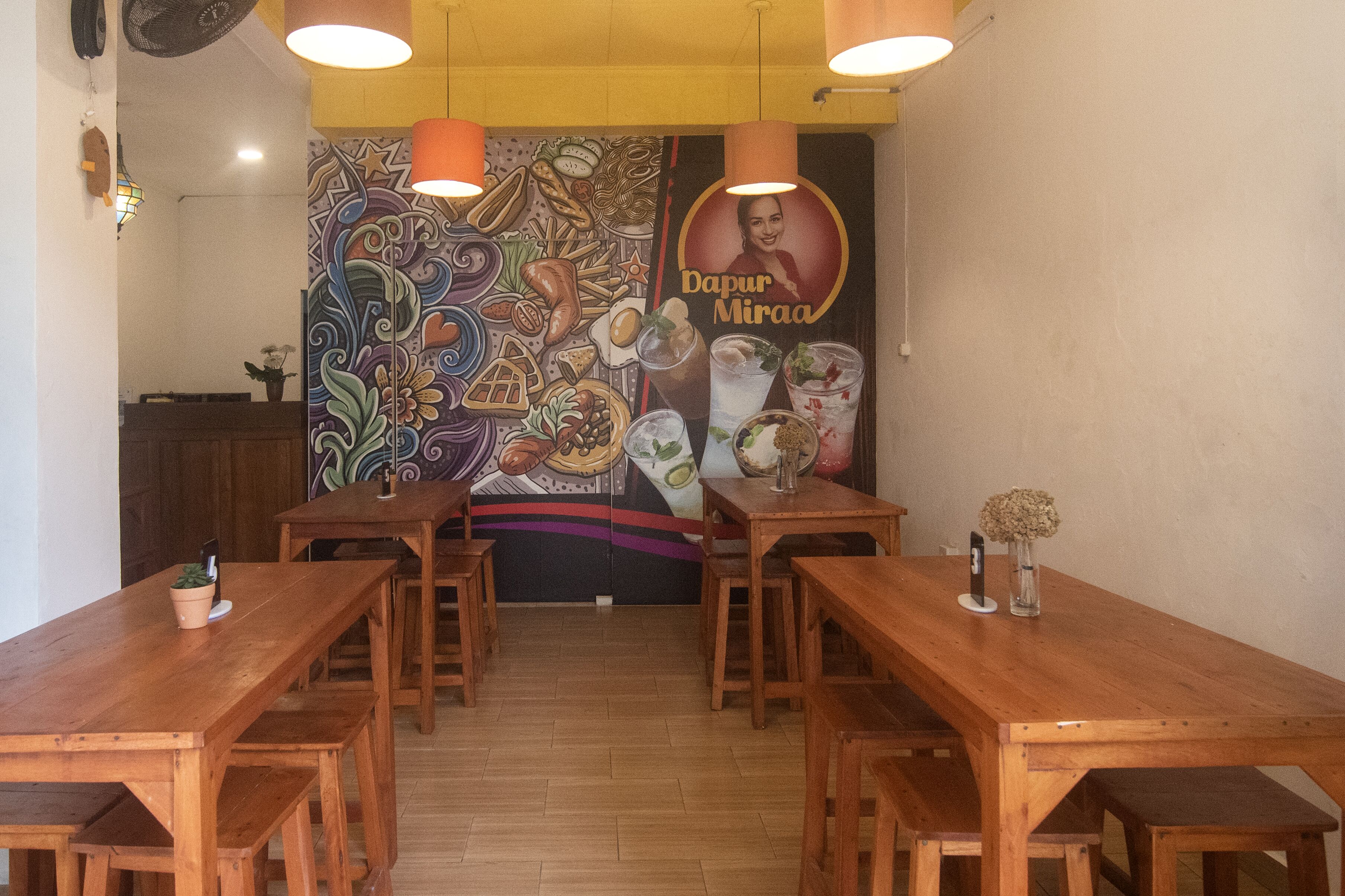 Ein Restaurant mit Holztischen und -bänken, einer Wandmalerei an der Rückwand und hängenden Pendelleuchten.