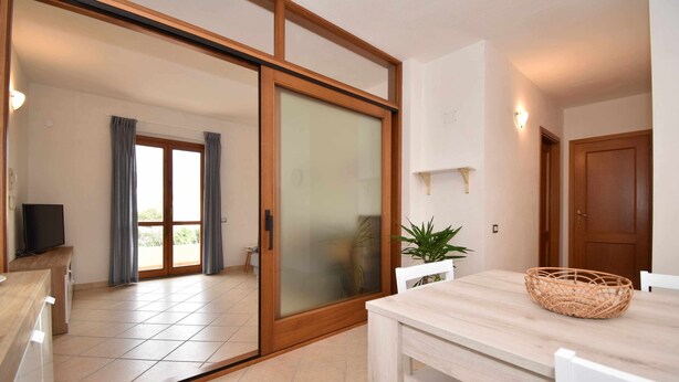 A modern room with a sliding glass door leading to a balcony, a wooden dining table, and a television on a stand. - Three-room apartment downtown with two bathrooms and A/C (Santa Teresa Gallura)