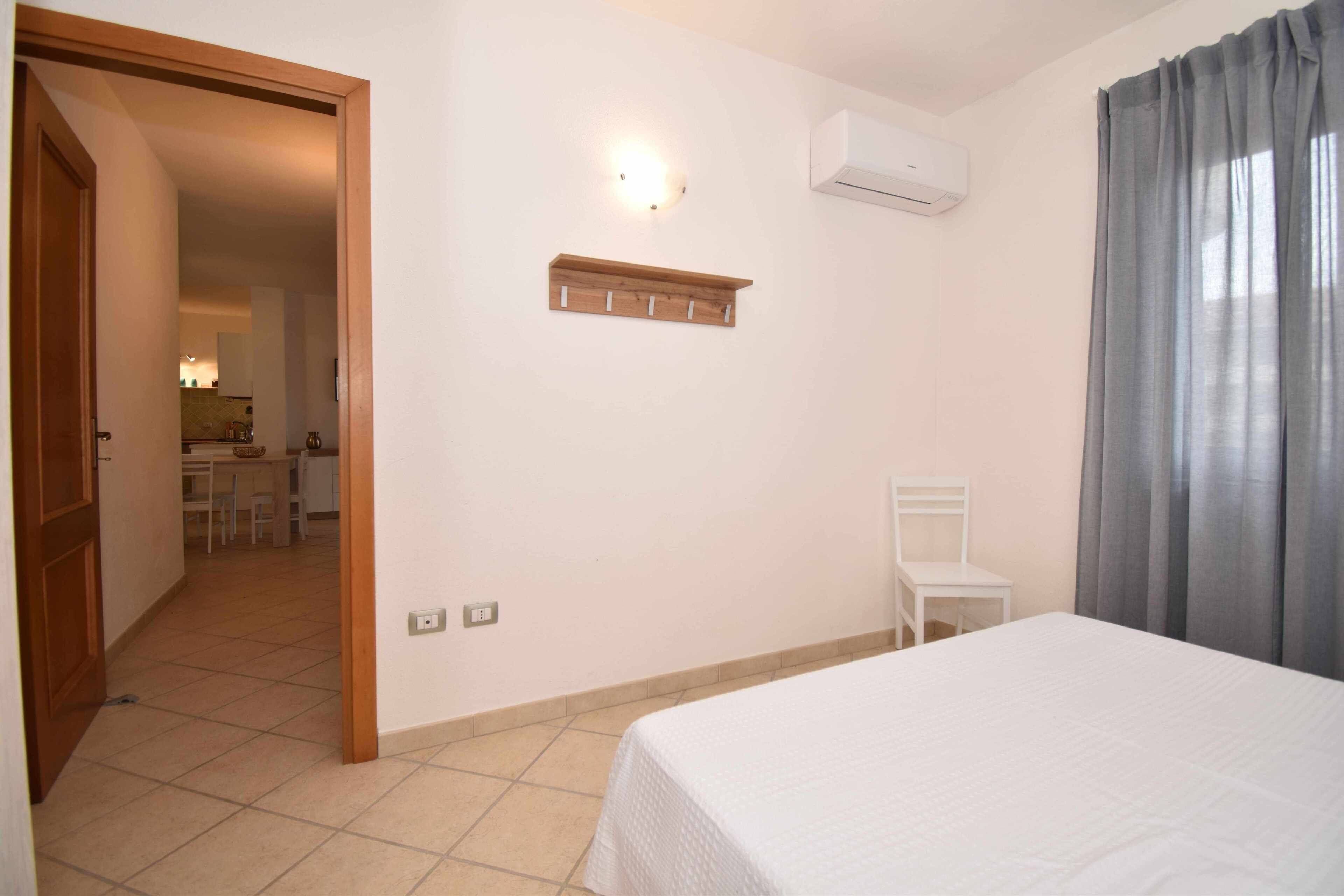 Une chambre d’hôtel avec un lit, une chaise, une petite table, une étagère fixée au mur et une salle de bain visible à travers une porte ouverte.